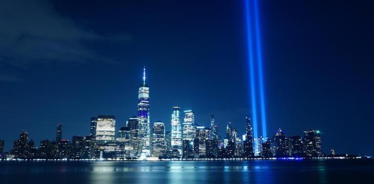 9/11 memorial of nighttime NYC skyline with two spotlights in place of the towers.